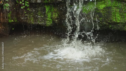 Closeup falling water into muddy pool, mosslined grotto with turbulent splash and rising sediment, wet stone ledge showing erosion, textured ripples and ambient damp atmosphere