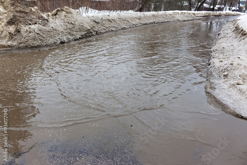 Melting snow creates a muddy water channel along the roadside, reflecting overcast spring weather and winter's retreat