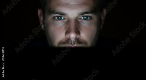 Man's intense gaze illuminated by the glow of a digital screen, reflecting deep concentration at night