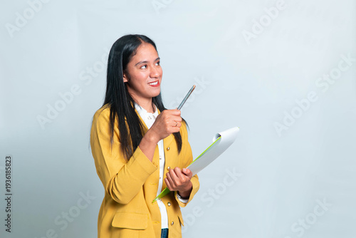 Smiling Indonesian woman wearing yellow blazer and white shirt, holding green clipboard and raising pen while looking up at copy space, isolated on white background.