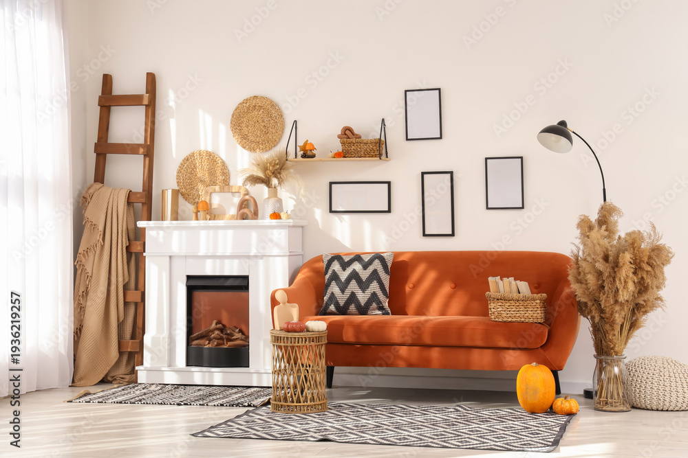 Naklejka premium Interior of living room with orange sofa, ladder, lamp, coffee table and autumn home decor on fireplace near white wall