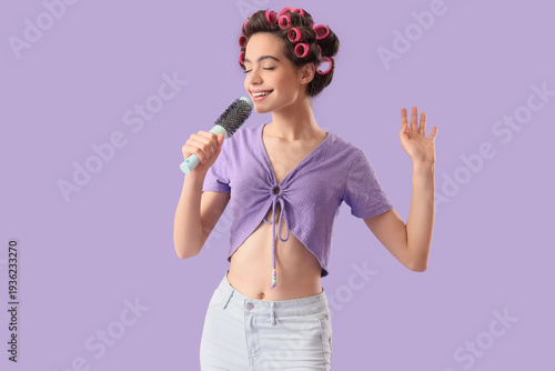 Young woman with hair curlers and brush singing on lilac background