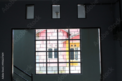 Interior view through a dark window frame with security bars looking out to colorful pink and yellow houses. Creative framing composition exploring the contrast between indoor shadow and bright outdoo