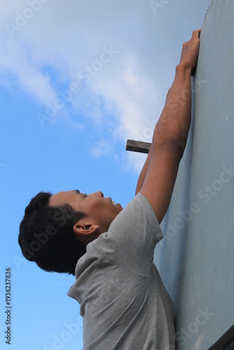 Side profile of a young man in a grey t-shirt climbing or hanging on a grey wall against a clear blue sky. Concept of effort, challenge, ambition, and reaching for goals. Dynamic low-angle shot showin