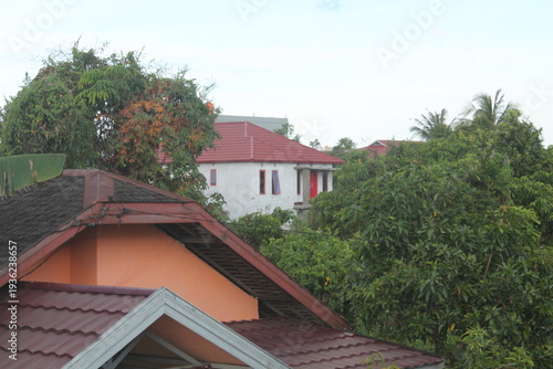 High angle view of residential houses with red clay tile roofs nestled among lush tropical greenery. Beautiful contrast between urban development and nature under a bright afternoon sky.