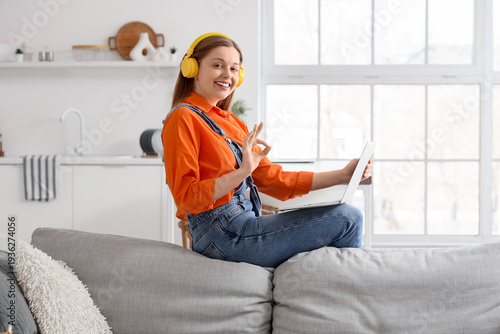 Smiling young woman in headphones using laptop on sofa at home and showing ok...