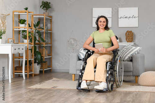 Beautiful young woman in wheelchair with headphones using tablet computer at ...