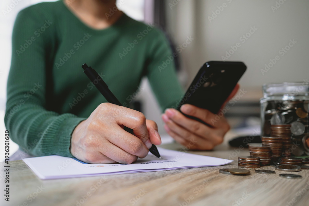 Fototapeta premium Woman writing budget notes while using smartphone with coins and savings jar on table. Representing personal finance, budgeting, saving money and financial planning.