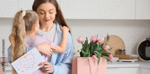 Cute little girl hugging her mom and greeting for Mother's Day with tulips and postcard in kitchen