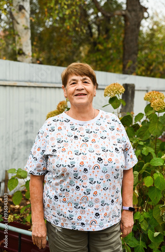Portrait of a 75 year old Russian woman smiling outdoors in a rural village garden with hydrangea plants on a cloudy autumn day.