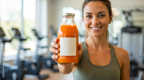 Smiling fitness woman holding bottle of fresh carrot juice in modern gym promoting natural nutrition healthy lifestyle and sports wellness