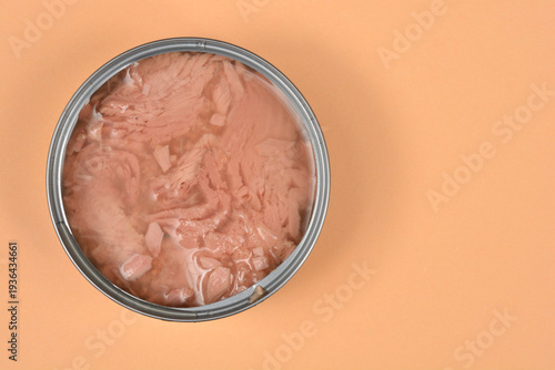 Top view of an open round metal can filled with tuna chunks in brine placed on an pale orange paper surface with copy space on the right