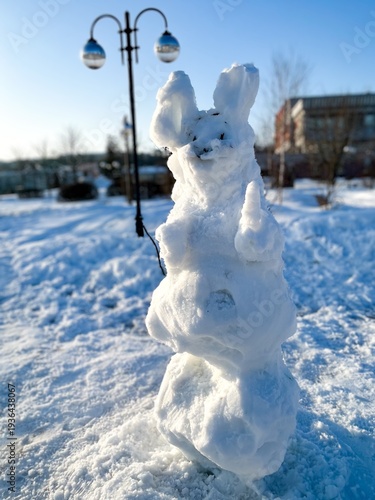 Snow sculpture resembling a rabbit standing next to a lamp post on a bright winter day. Frosty landscape with snow-covered ground and trees in the background.
