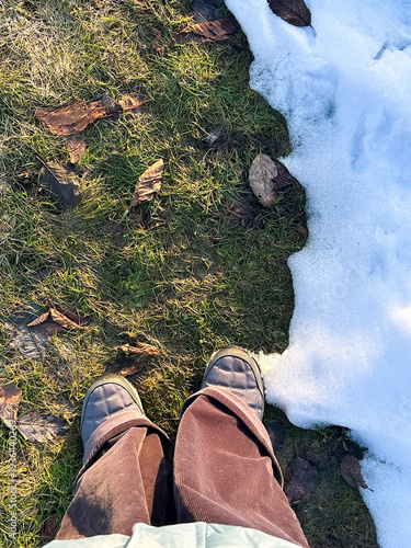 Feet stand on the dividing line between melting snow and new grass, symbolizing the transition from winter to spring. Brown leaves scattered on the grass indicate seasonal change.
