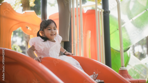 Happy little girl playing on an orange slide at an outdoor playground in warm sunlight, candid childhood fun and leisure concept.