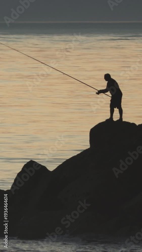 A man stands on rocks and fishes in the sea at sunset