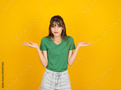 Photos Young woman shrugging shoulders with spread arms, looking at camera with unsure face expression, isolated on yellow background