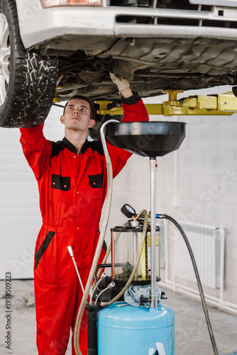 Professional auto mechanic in red uniform working under a car on a lift in a service station. Technician performing oil change maintenance using specialized drainage equipment in a modern workshop gar