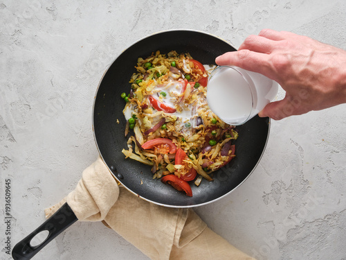 Cooking a vegetable stir-fry with rice in a pan while adding milk in a kitchen setting