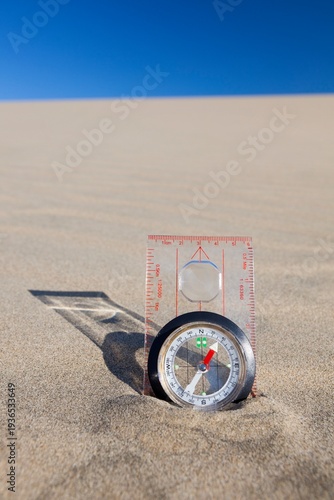 Orienteering compass placed upright in a sand dune beneath a clear blue sky, symbolizing navigation and exploration.