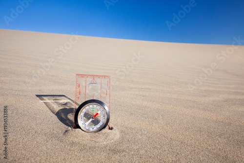 Orienteering compass placed upright in a sand dune beneath a clear blue sky, symbolizing navigation and exploration.