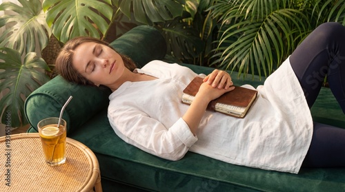Peaceful woman sleeping on a green velvet sofa with a book surrounded by lush plants.