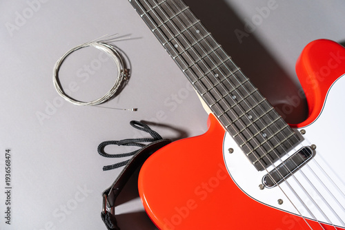 Close-up of a red electric guitar with white pickguard, dark fretboard, metal frets, dot inlays, coiled replacement strings, and a black strap with a leather end placed beside the neck.