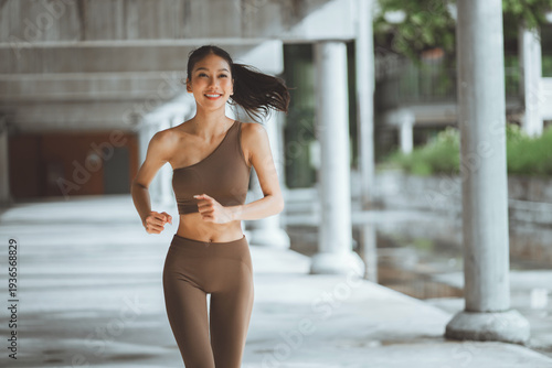 A beautiful and fit young Southeast Asian woman wearing a brown sports bra, running and smiling with confidence through urban park during her morning workout, representing a healthy lifestyle