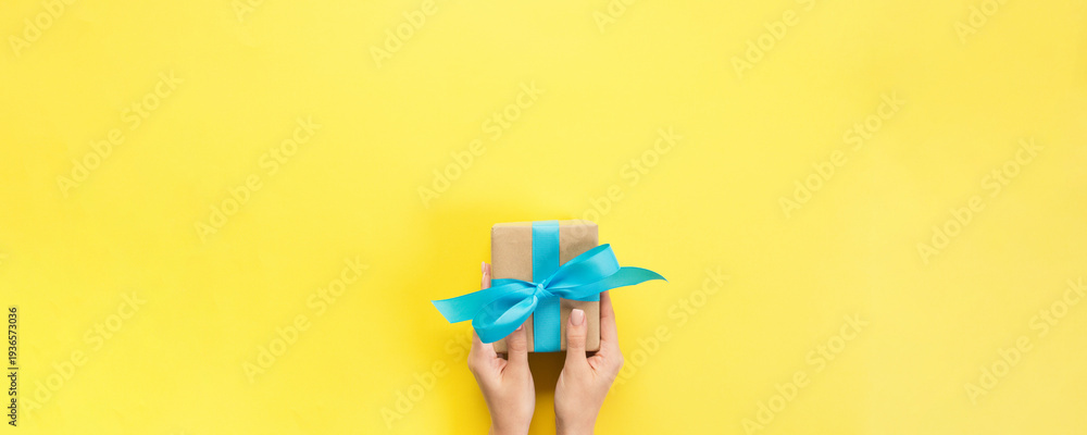 Fototapeta premium Woman arms holding gift box with blue ribbon on color background, top view