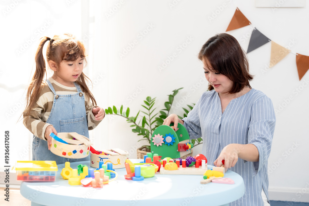 Naklejka premium Asian single mother playing with kids at home, mom teaching preschool daughter lacing activity, toddler son watches, home daycare learning environment, positive parenting supports teamwork indoors