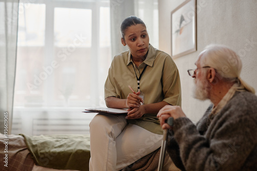 Young adult Black woman social worker sitting on bed, holding clipboard, talking to senior Caucasian man with long hair and beard using walking cane in residential setting