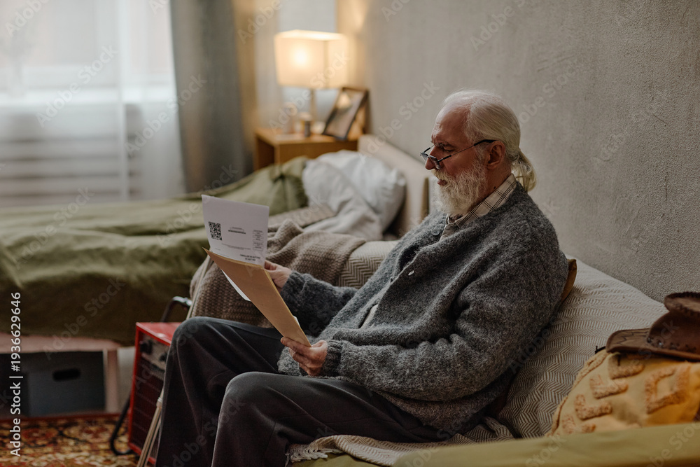 Fototapeta premium Senior Caucasian man sitting on bed checking utility bills with glasses, holding paper in both hands, appearing focused, gray hair and beard visible, bedroom setting with soft lighting