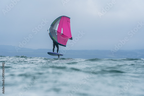 A man wing foils at sea using a pink and grey hand held inflatable wing, riding a hydrofoil surfboard. Green sea, cloudy blue sky.	
