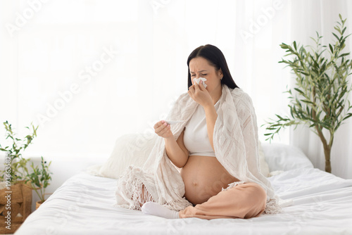 Schilderij op canvas Pregnant woman sitting on bed wrapped in blanket checking thermometer while feeling unwell