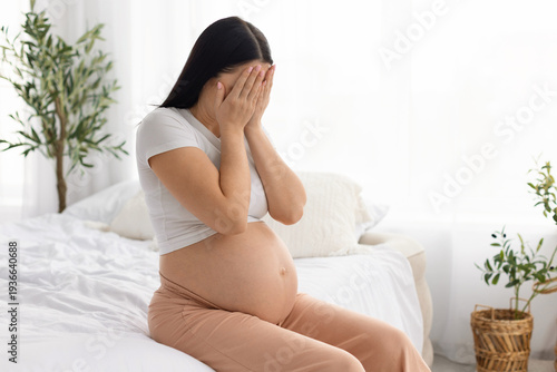 Fotografie Pregnant woman sitting on bed covering face feeling emotional stress during pregnancy