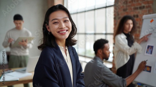 Confident creative lead pausing to share an authentic, proud smile with the viewer while her design team actively maps out visual concepts on a whiteboard behind her.