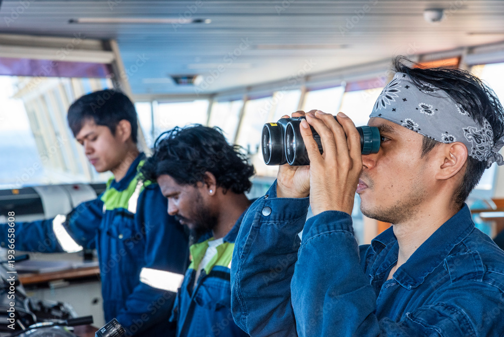 Obraz premium Professional ship crew focused on bridge control systems as one deck officer conducts visual lookout with binoculars to ensure safe navigation at sea.