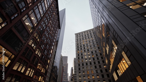 dusk city street framed by skyscrapers, warm office lights glow inside brick and glass facades, reflective surfaces mirror neighboring towers, narrow canyon