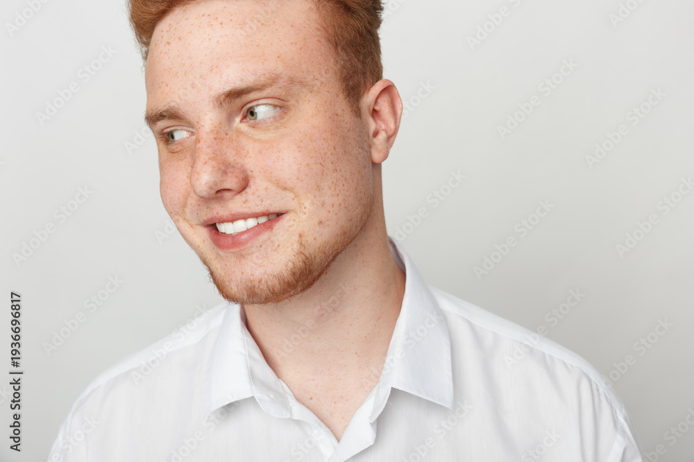 Obraz premium Young redhead man with freckles smiles looking sideways wearing white shirt on light background. Concept of casual style, happiness, and natural male portrait in studio lighting.