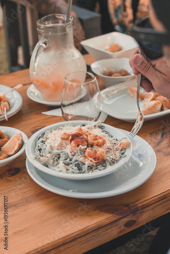 Primer plano de persona comiendo pasta con camarones en una mesa de restaurante al aire libre con bebidas y entradas