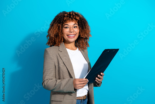 Εκτύπωση καμβά Confident young woman in a plaid blazer holds a clipboard smiling against blue b