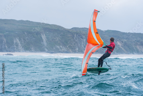 A man wing foils at sea using a bright orange hand held inflatable wing, riding a hydrofoil surfboard. Green sea, cloudy blue sky.	 Coastal cliffs in the background.