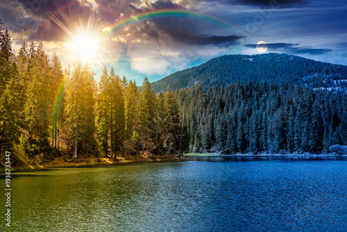 mountain lake among the green spruce forest in beautiful spring landscape. day meet night. sun and moon over rippled water surface. alpine nature scenery with clouds on sky over the evergreen woods