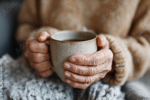 Warm hands cradle a ceramic mug. Soft knuckles hint at gentle age. Cozy sweater wraps around a quiet moment. Cuddling blanket adds comfort to the scene. Stillness speaks of peace and quiet reflection