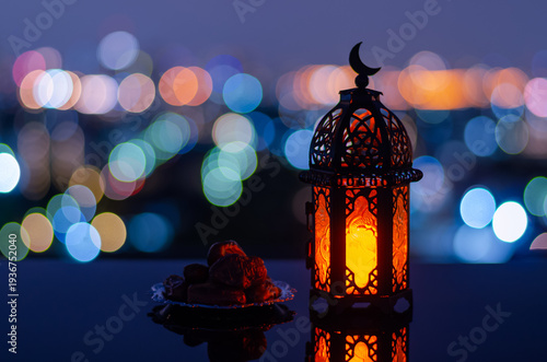 Selective focus of lantern that have moon symbol on top and small plate of dates fruit with city bokeh light background for the Muslim feast of the holy month of Ramadan Kareem.