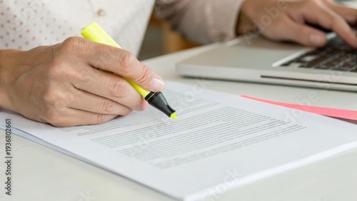 A person highlights text on a document with a yellow marker while using a laptop, suggesting multitasking or reviewing work.
