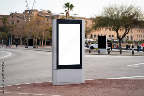 Digital street kiosk displaying a blank white screen, offering copy space for advertising at a Barcelona city intersection, with blurry urban background