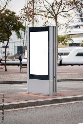 Outdoor blank billboard advertising mockup on a city street, displaying a vertical white screen for promotional messages in an urban environment with trees and a parked yacht