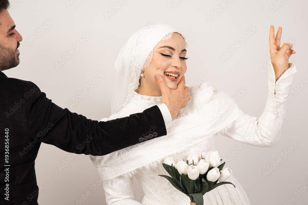 Naklejka premium Playful studio portrait of a happy bride and groom sharing a joyful moment on their wedding day. The groom gently holds the bride’s chin while she smiles and makes a peace sign gesture