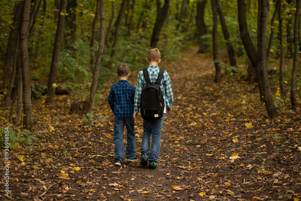 Obraz premium Two boys walking on a forest path in autumn, wearing casual clothes and backpacks, surrounded by trees and fallen leaves.